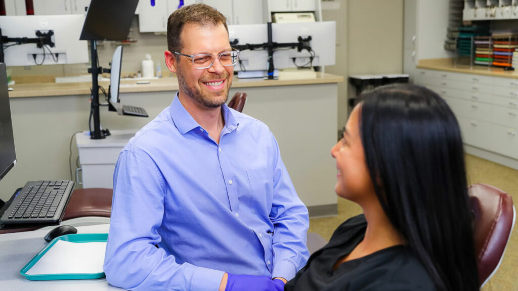 What is an orthodontist? A dentist, Dr. Warren Libby, DDS, wearing glasses and a blue shirt is smiling and talking to a patient with long black hair in a dental office.